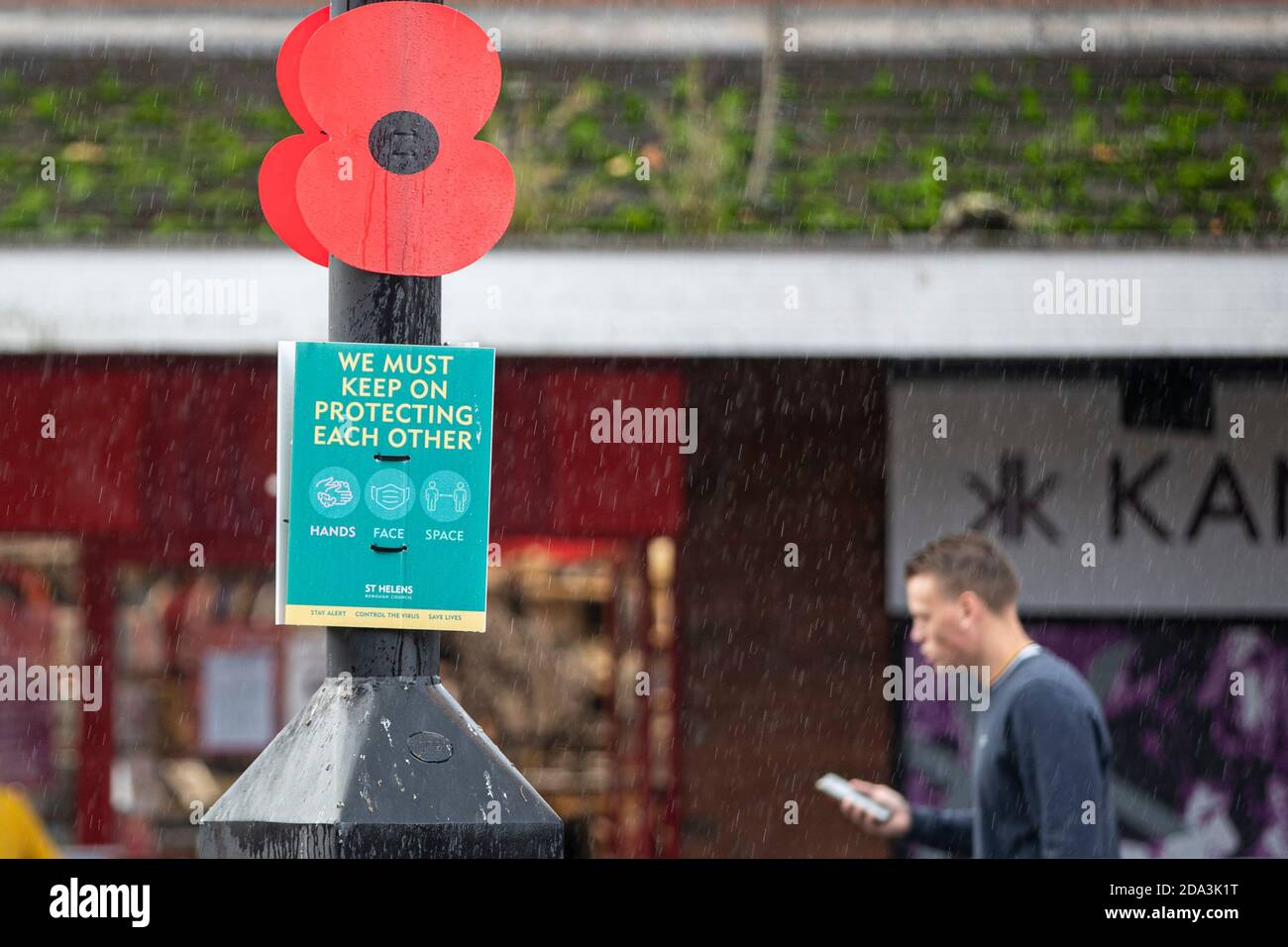 © concesso in licenza a London News Pictures. 03/11/2020. St Helens, Regno Unito. Un uomo cammina da un papavero per ricordare accanto a un segno covid mentre la gente ha coraggioso la pioggia dentro Foto Stock