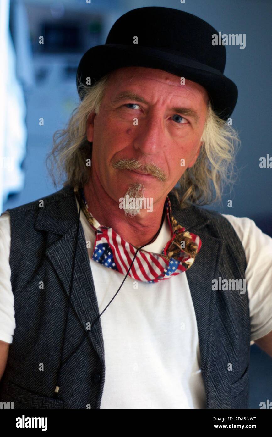 Ritratto di bell'uomo biondo dagli occhi azzurri con cappello bowler e  gilet con bandiera americana con maschera coved-19 intorno al collo.  Guardando al fianco con i capelli lunghi, mu Foto stock -