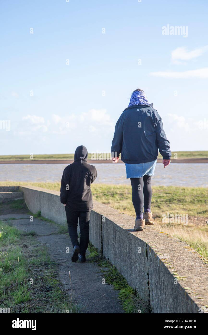 Madre e figlio adulto che camminano insieme su una parete bassa vicino al fiume ore, Suffolk, Inghilterra, Regno Unito Foto Stock