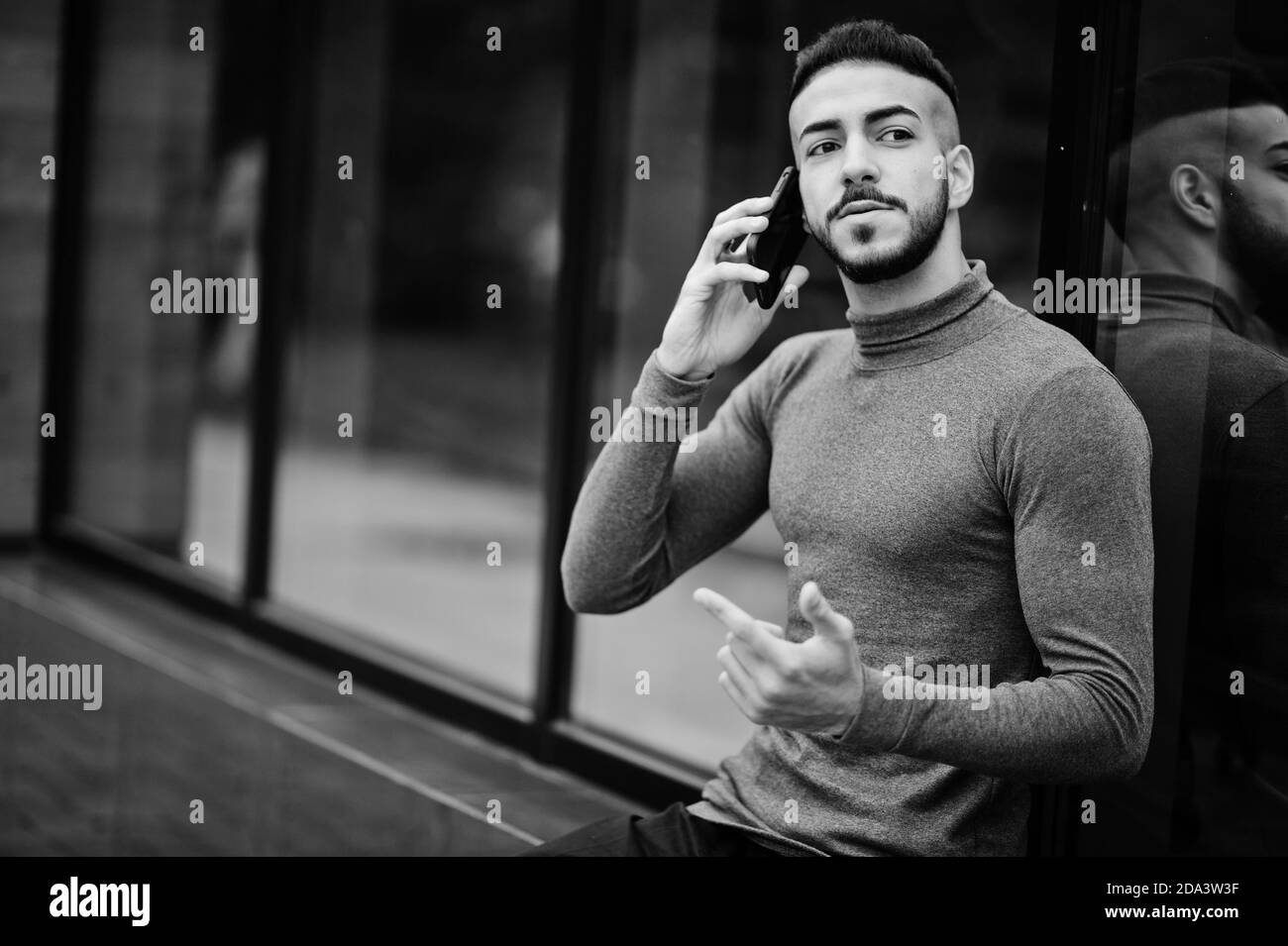 Ritratto di elegante barba araba uomo indossare dolcevita grigio. Modello arabo ragazzo sedersi e parlare con smartphone. Foto Stock