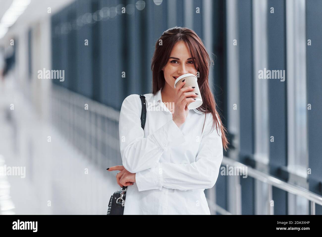 Brunette in camicia bianca all'interno in un moderno aeroporto o corridoio durante il giorno e tenendo una tazza di bevanda Foto Stock