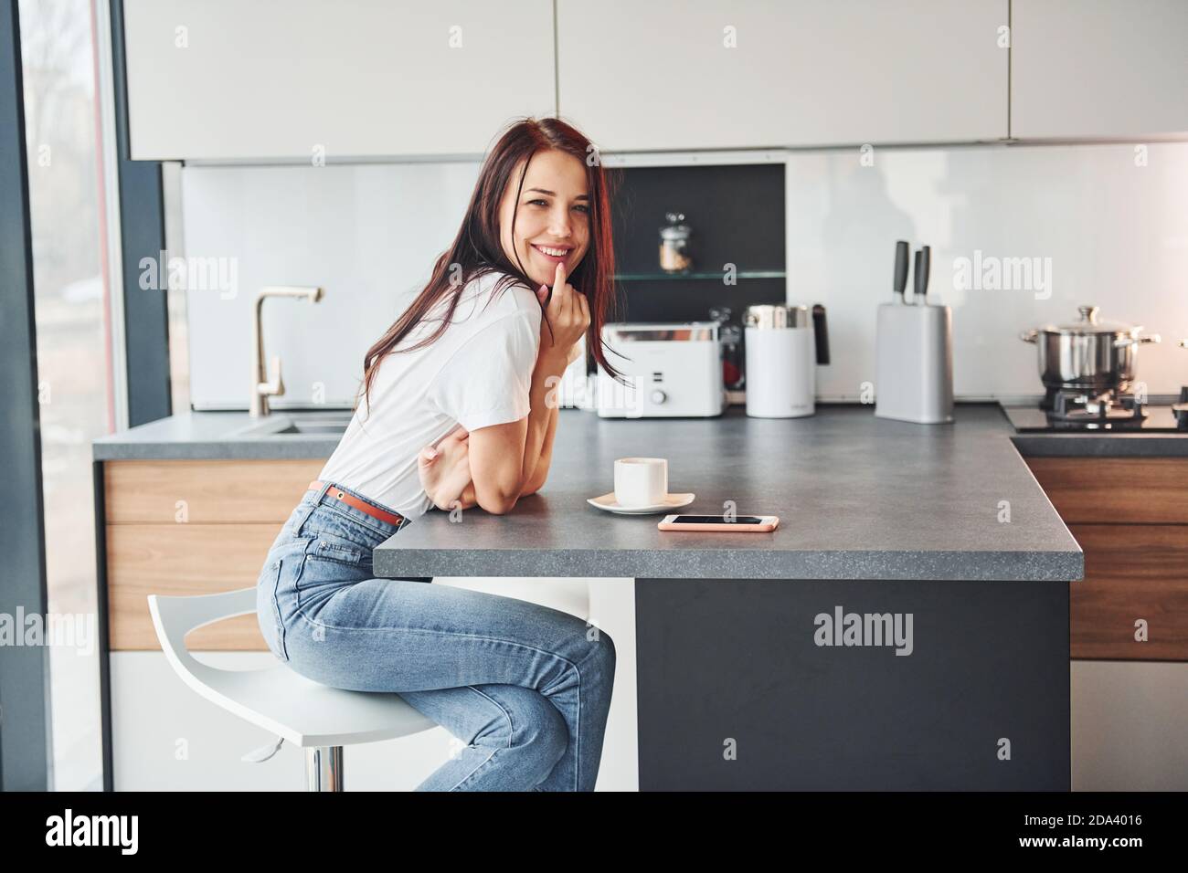 Giovane bella brunetta in abiti casual si siede in cucina con bevanda fresca al mattino Foto Stock