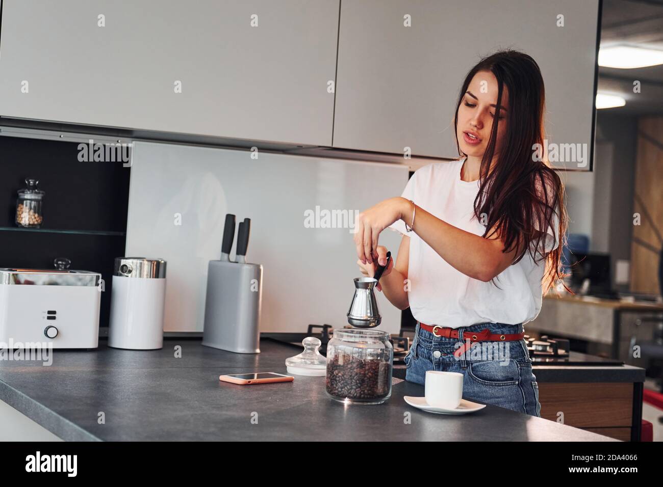 Giovane bella brunetta in abiti casual preparare caffè in cucina di giorno Foto Stock