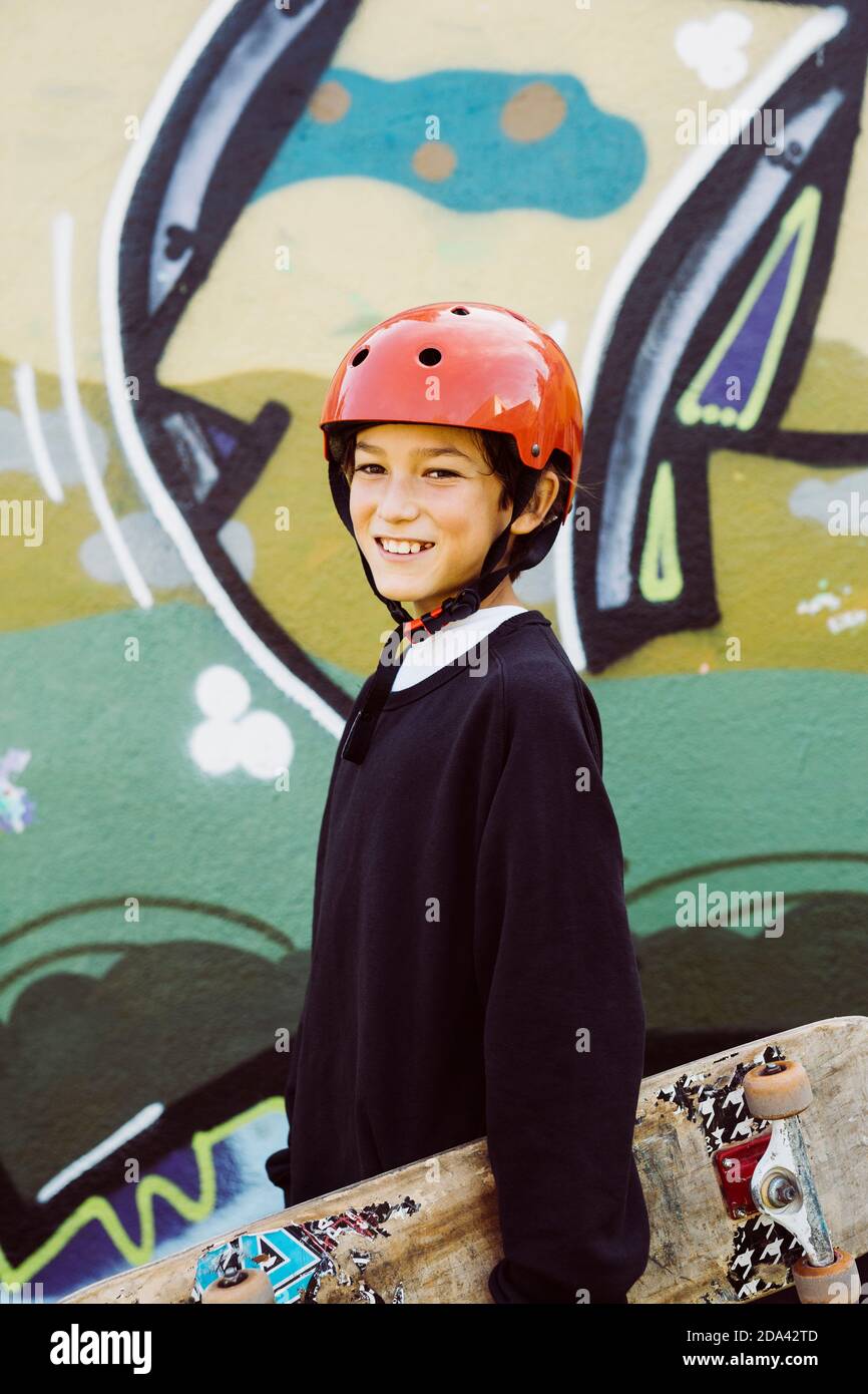 Ritratto di un giovane skater con un vecchio skateboard e un casco rosso davanti a un murale di graffiti Foto Stock