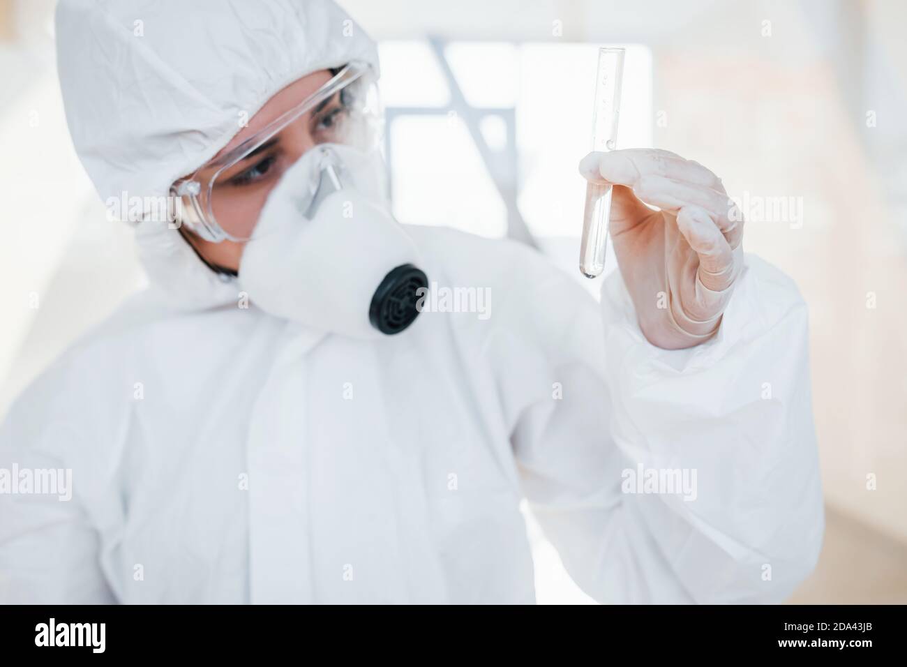 Con provetta in mano. Dottoressa scienziata in camice da laboratorio, occhiali difensivi e maschera in piedi in interni Foto Stock