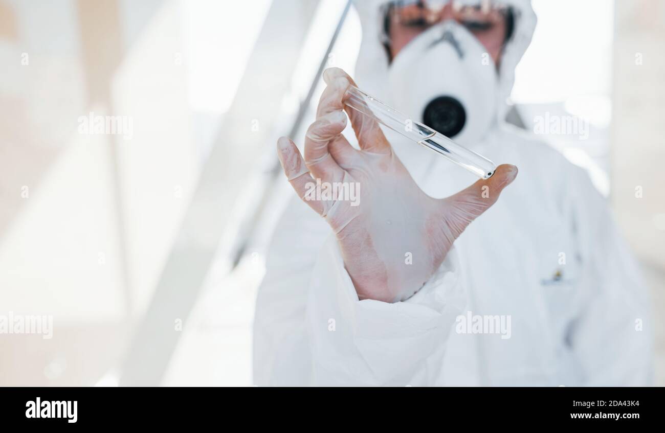 Con provetta in mano. Dottoressa scienziata in camice da laboratorio, occhiali difensivi e maschera in piedi in interni Foto Stock