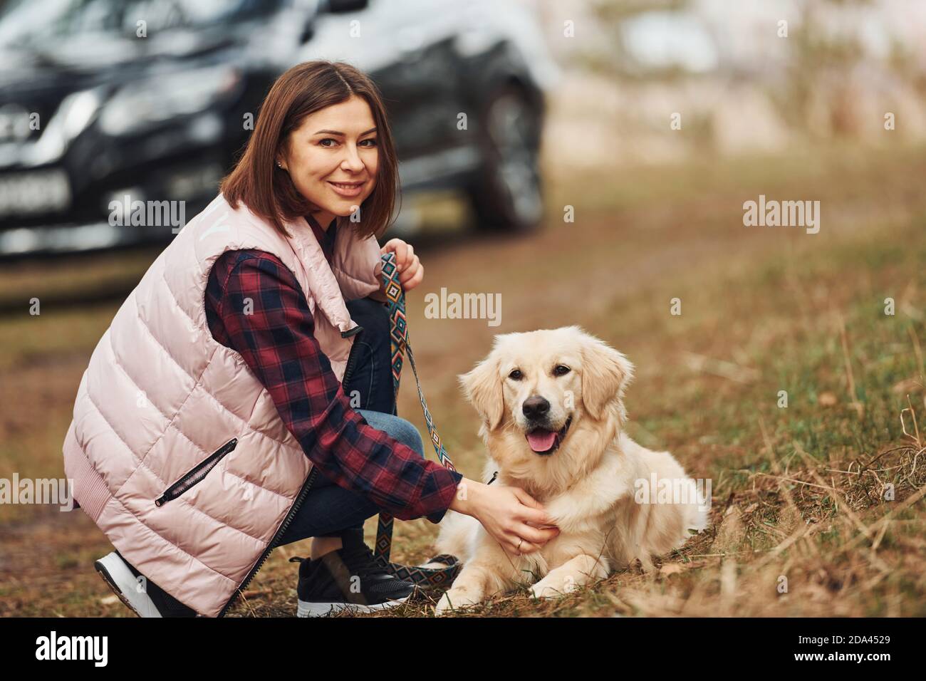 Donna in abiti caldi seduta con il suo cane in foresta contro la moderna auto nera Foto Stock