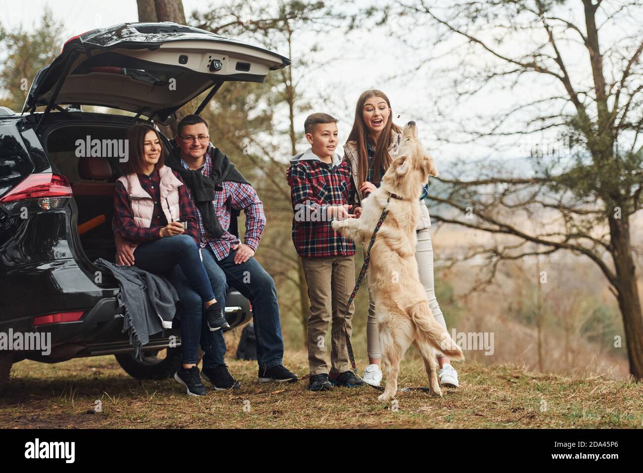 La famiglia felice si diverte con il loro cane vicino a un'auto moderna all'aperto nella foresta Foto Stock