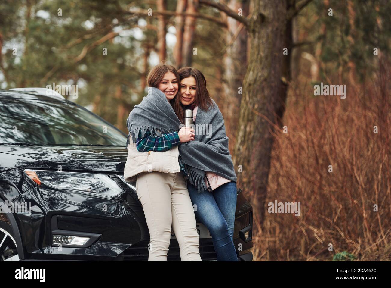 Madre e figlia si levano in piedi insieme vicino all'auto nera moderna all'aperto nella foresta Foto Stock