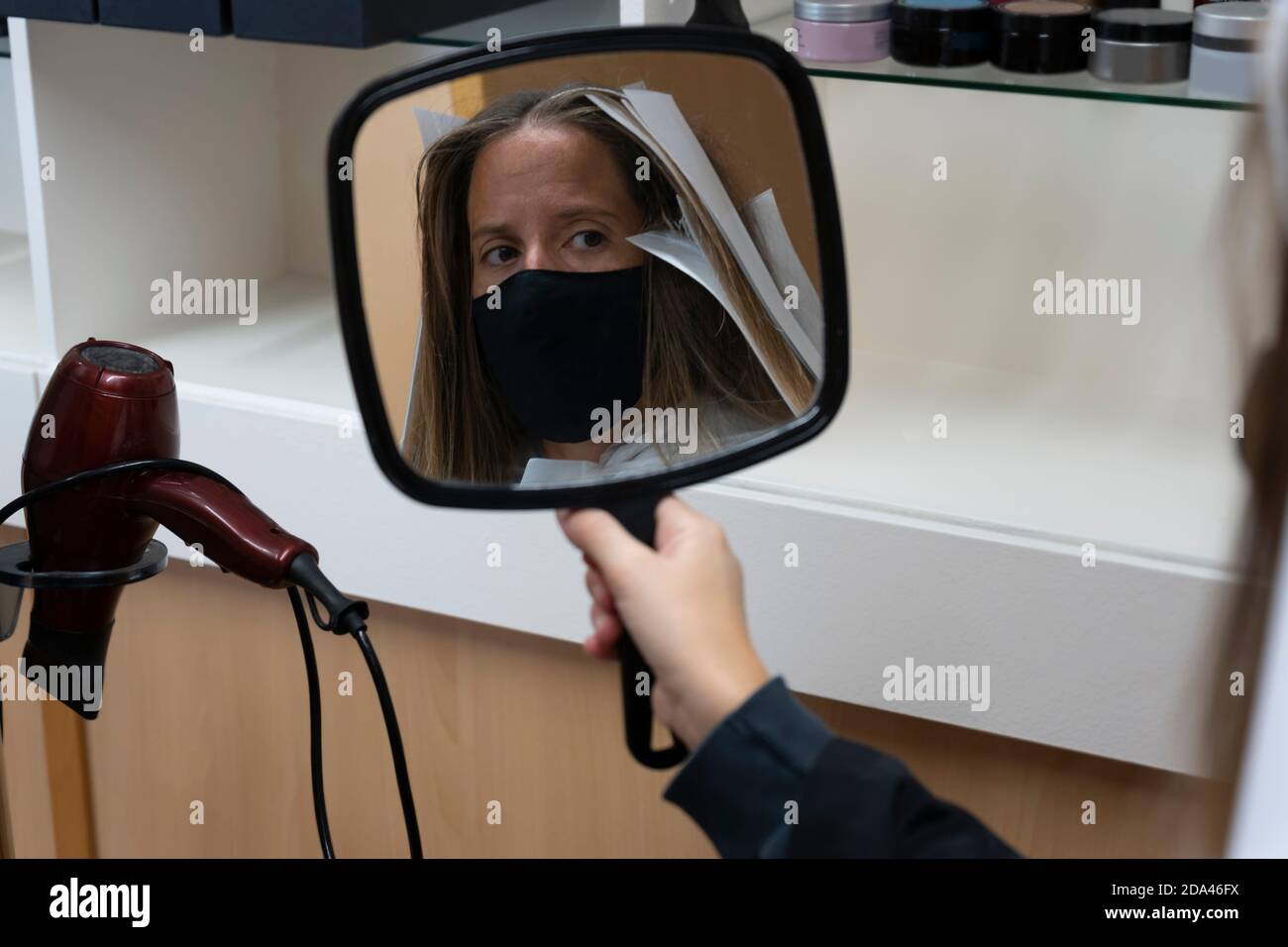 Cliente donna con maschera medica protettiva in parrucchiere, guardando se stessa in mano specchio il trattamento.Coronavirus covid-19 concetto. Foto Stock
