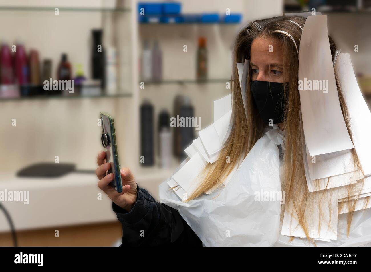 Bella donna cliente in parrucchiere utilizzando smartphone, prendendo un selfie , indossare maschera medica contro il coronavirus e carta tintura capelli per colorina Foto Stock