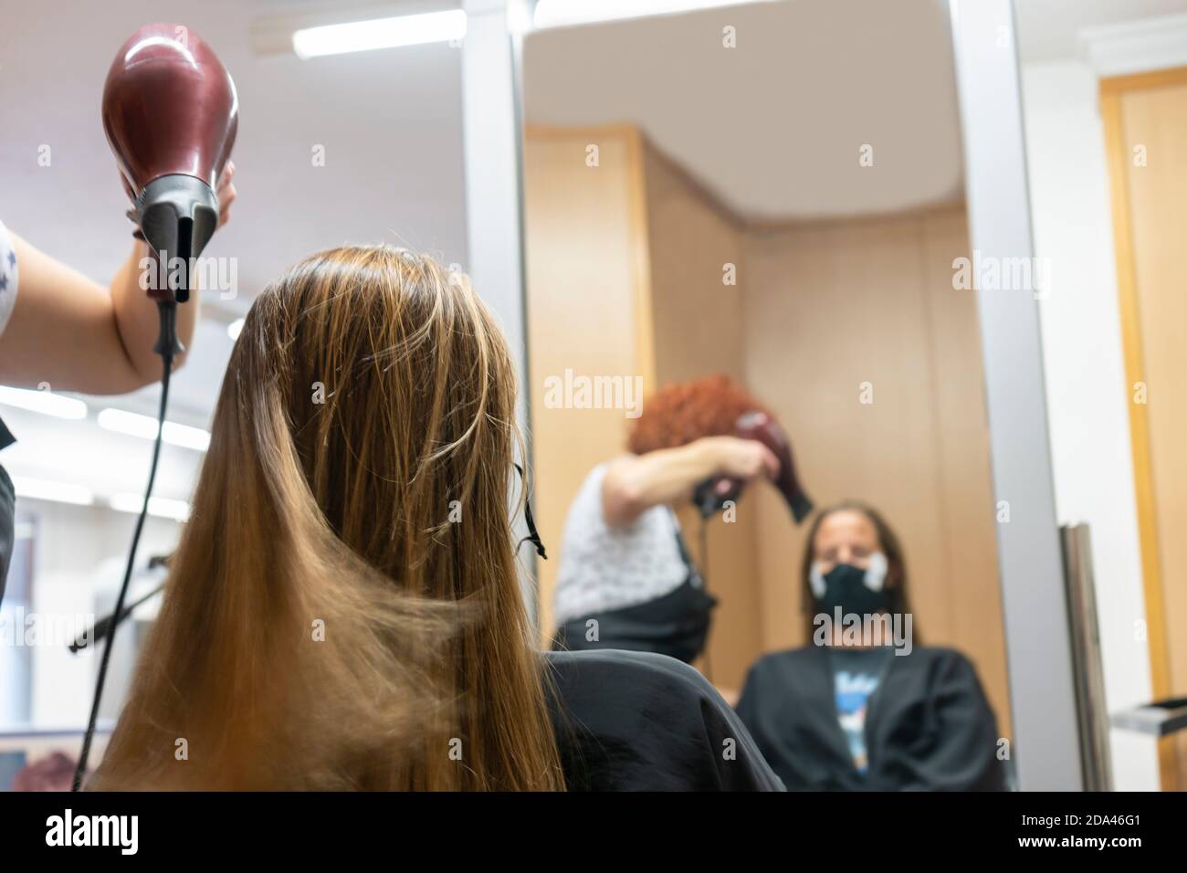 Professionale parrucchiere, stilista che fa acconciatura, asciugando e soffiando i capelli con un asciugacapelli in salone di bellezza, riflesso in uno specchio.indossare maschera medica Foto Stock