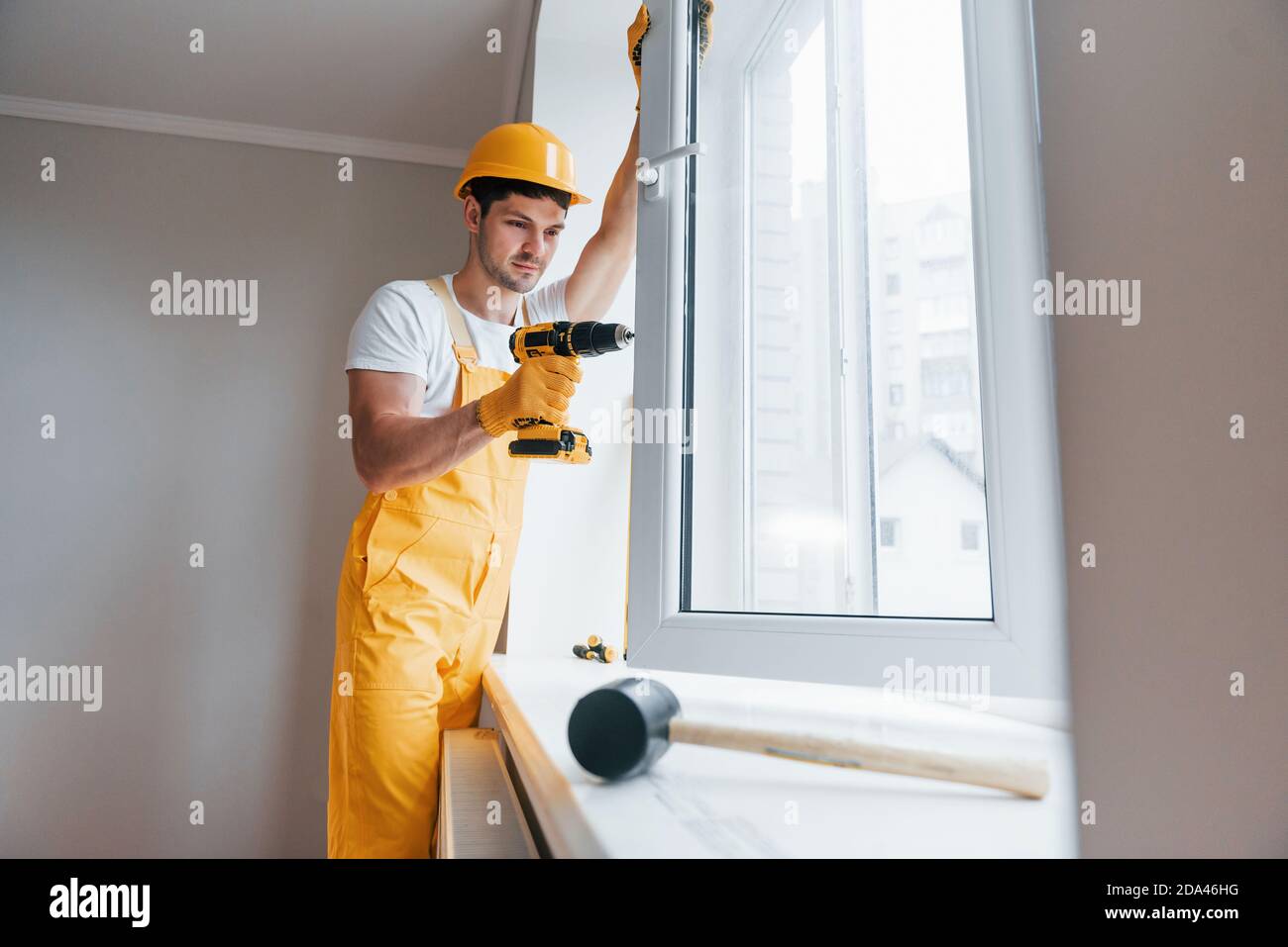 Handyman in uniforme gialla installa una nuova finestra utilizzando un cacciavite automatico. Ristrutturazione casa concezione Foto Stock