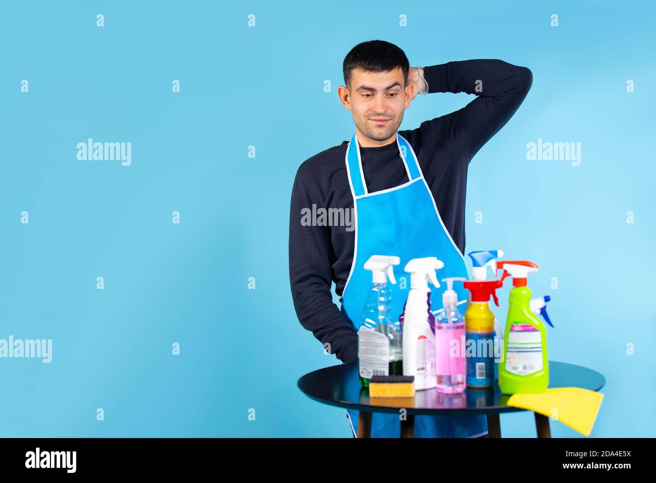 Stereotipi di genere. L'uomo tiene la testa e guarda il set di prodotti per la pulizia. Foto su sfondo blu con spazio laterale vuoto. Foto Stock