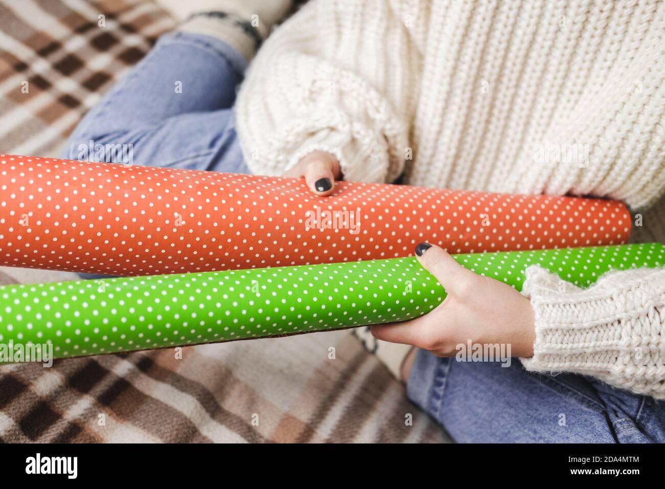 Giovane donna si siede su plaid in maglia accogliente pullover bianco lana, calzini e avvolge regalo di Natale in polka dot wrapping carta. Vassoio in legno con tazza di co Foto Stock