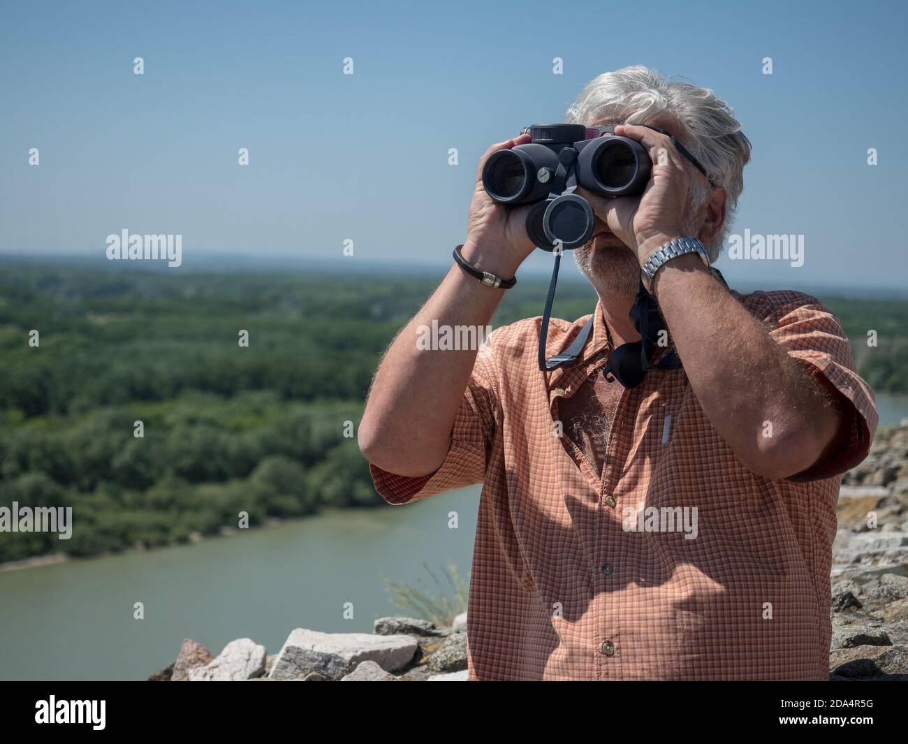 Uomo caucasico senior che guarda attraverso binocoli e osserva il paesaggio. Sfondo sfocato. Foto Stock