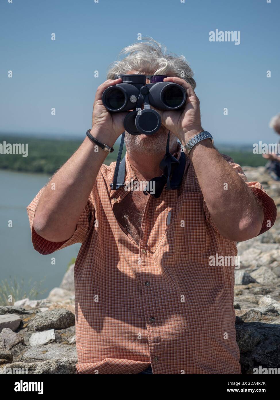 Uomo caucasico senior che guarda attraverso binocoli e osserva il paesaggio. Sfondo sfocato. Foto Stock