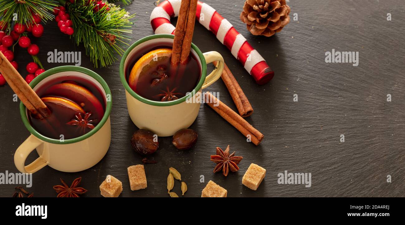 VIN brulé, bevanda alcolica invernale calda, due tazze d'epoca, ingredienti arancio e spezie e decorazioni natalizie su pietra nera, vista closeup. Foto Stock