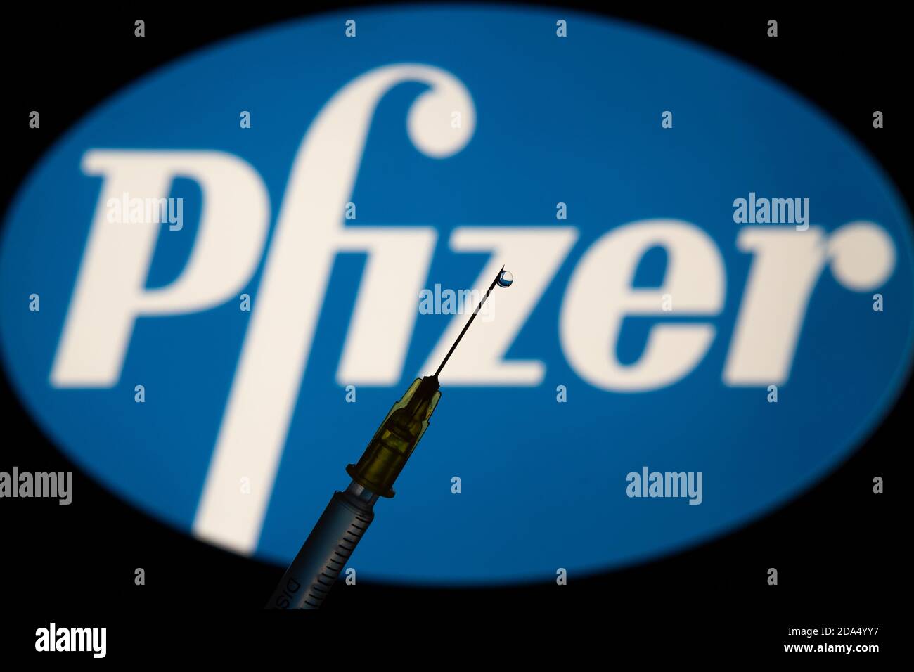 Stafford / Regno Unito - 9 novembre 2020: Concetto di vaccino Pfizer Covid-19. Ago della siringa e una goccia di liquido su di esso, logo della ditta di Pfizer sfocato sulla t Foto Stock