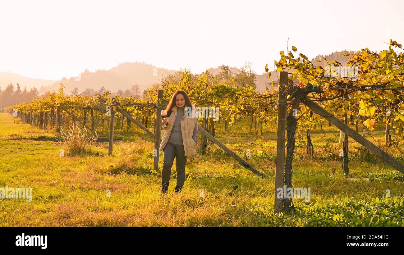 Tramonto sulle colline dell'Emmilia Romagna in autunno. Donna con una bottiglia di vino e un bicchiere in un'azienda agricola in italia. Abbagliamento del sole nel telaio. Banner, formato lungo. Spazio di copia. Foto di alta qualità Foto Stock