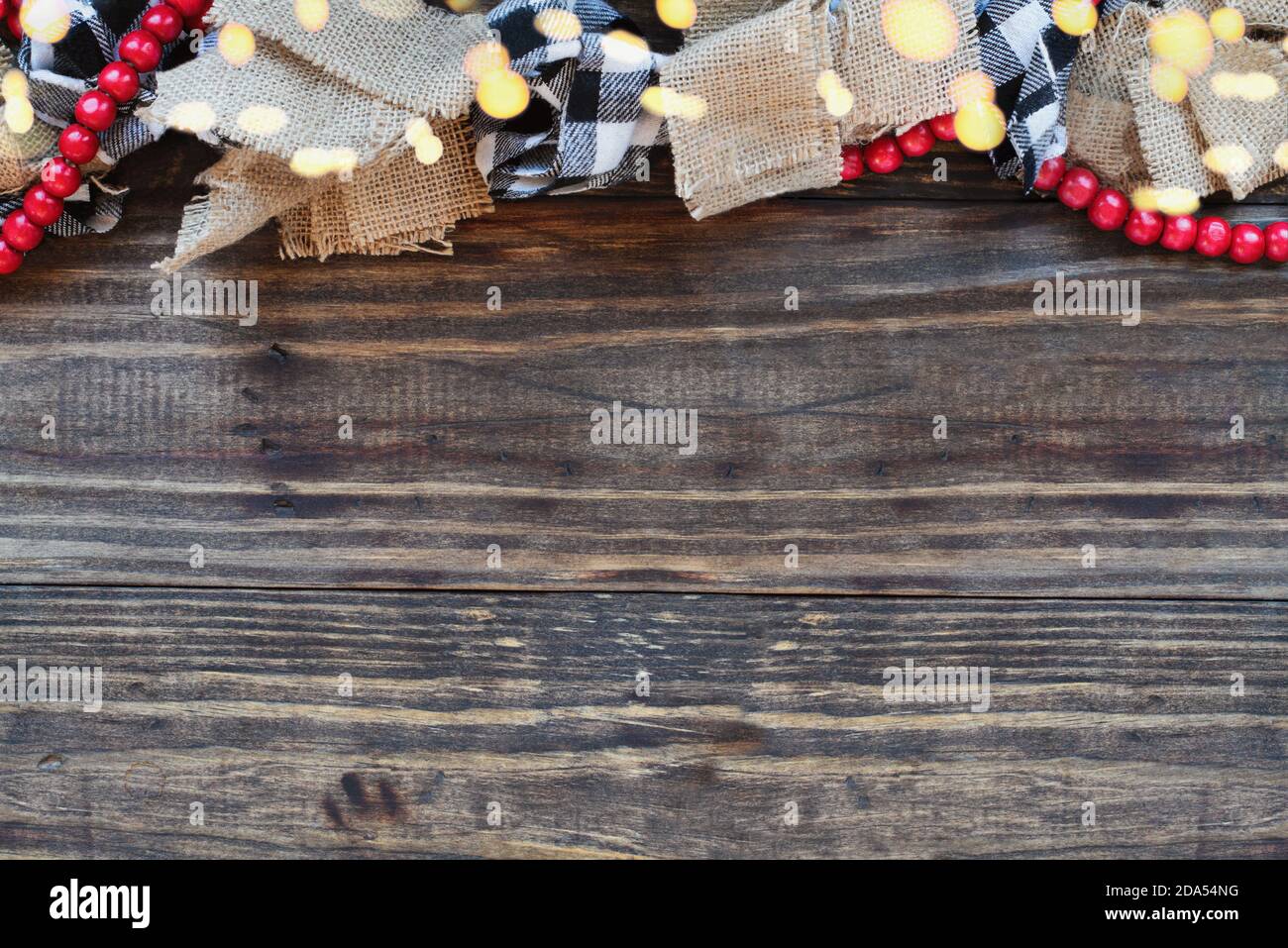 Ghirlanda di Natale con luci bokeh, burlap, tessuto bianco e nero di controllo bufalo e una fila di perle rosse su sfondo tavola di legno ruggine. Vista dall'alto Foto Stock