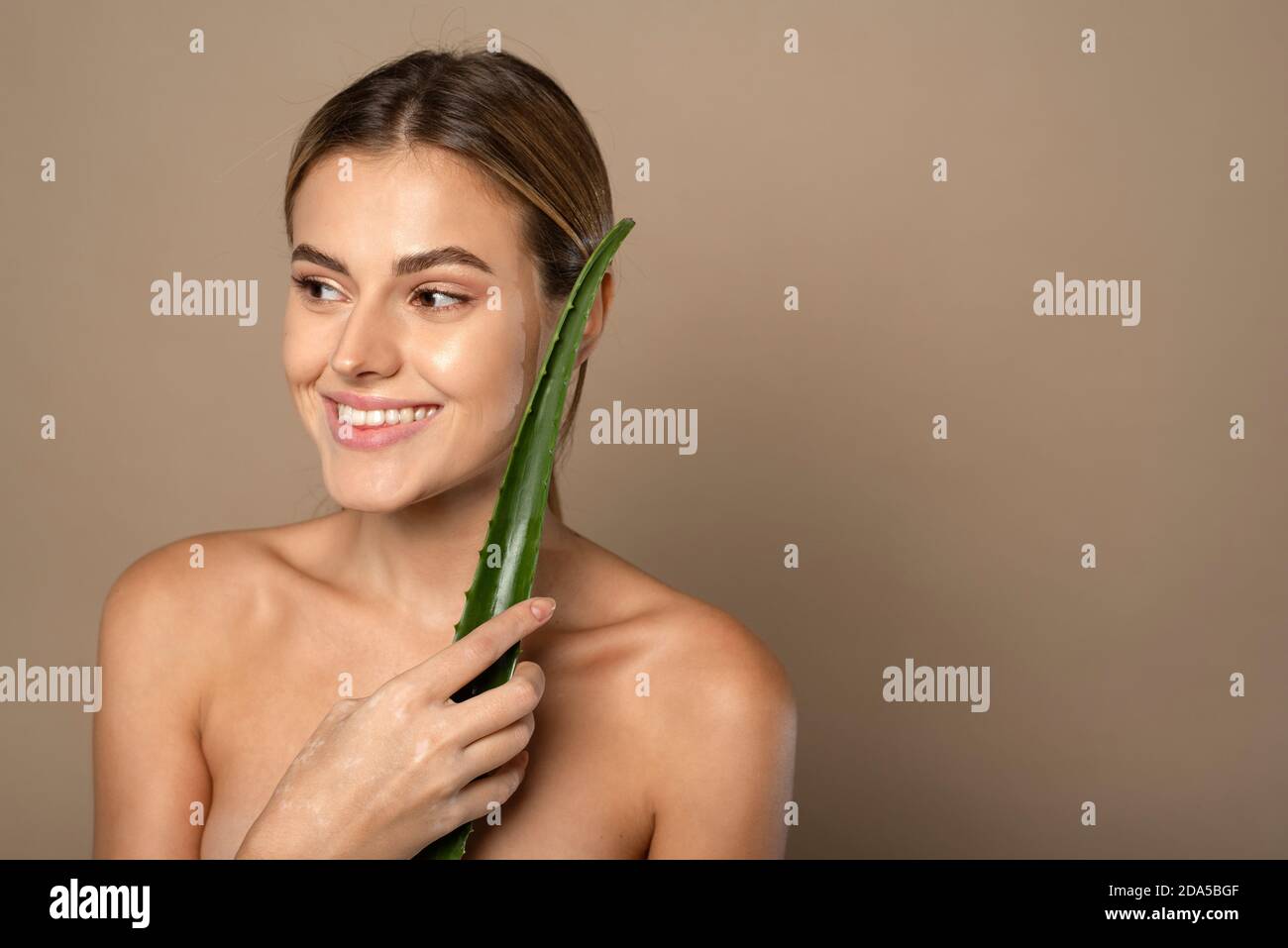 Sorridente giovane donna che tiene la foglia di aloe in mano su sfondo beige. Il concetto di cura della pelle, idratante con cosmetici naturali. Foto Stock