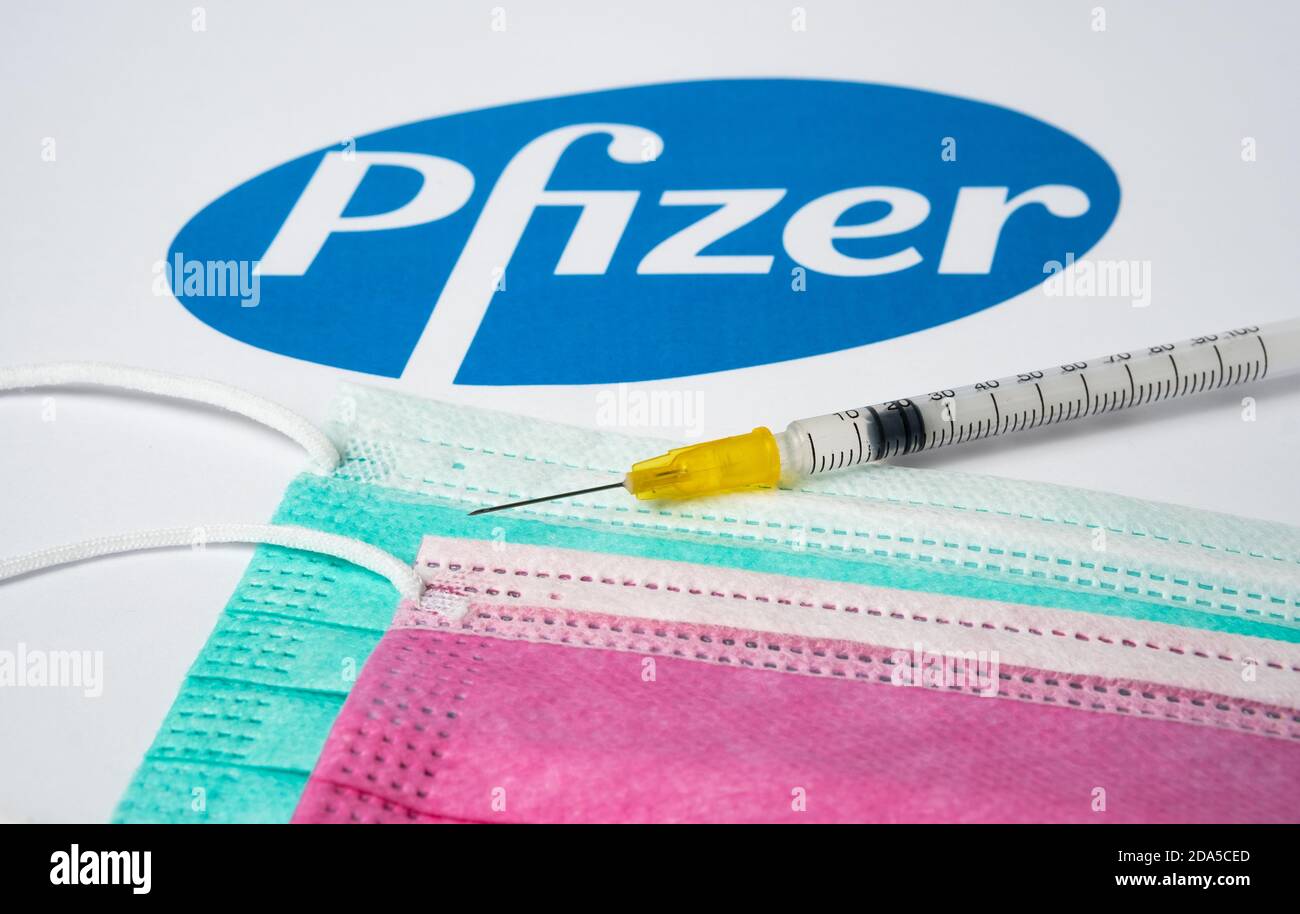 Stafford / Regno Unito - 9 novembre 2020: Concetto di vaccino Pfizer Covid-19. Siringa posizionata su maschere mediche e logo aziendali sfocati sulla ba Foto Stock