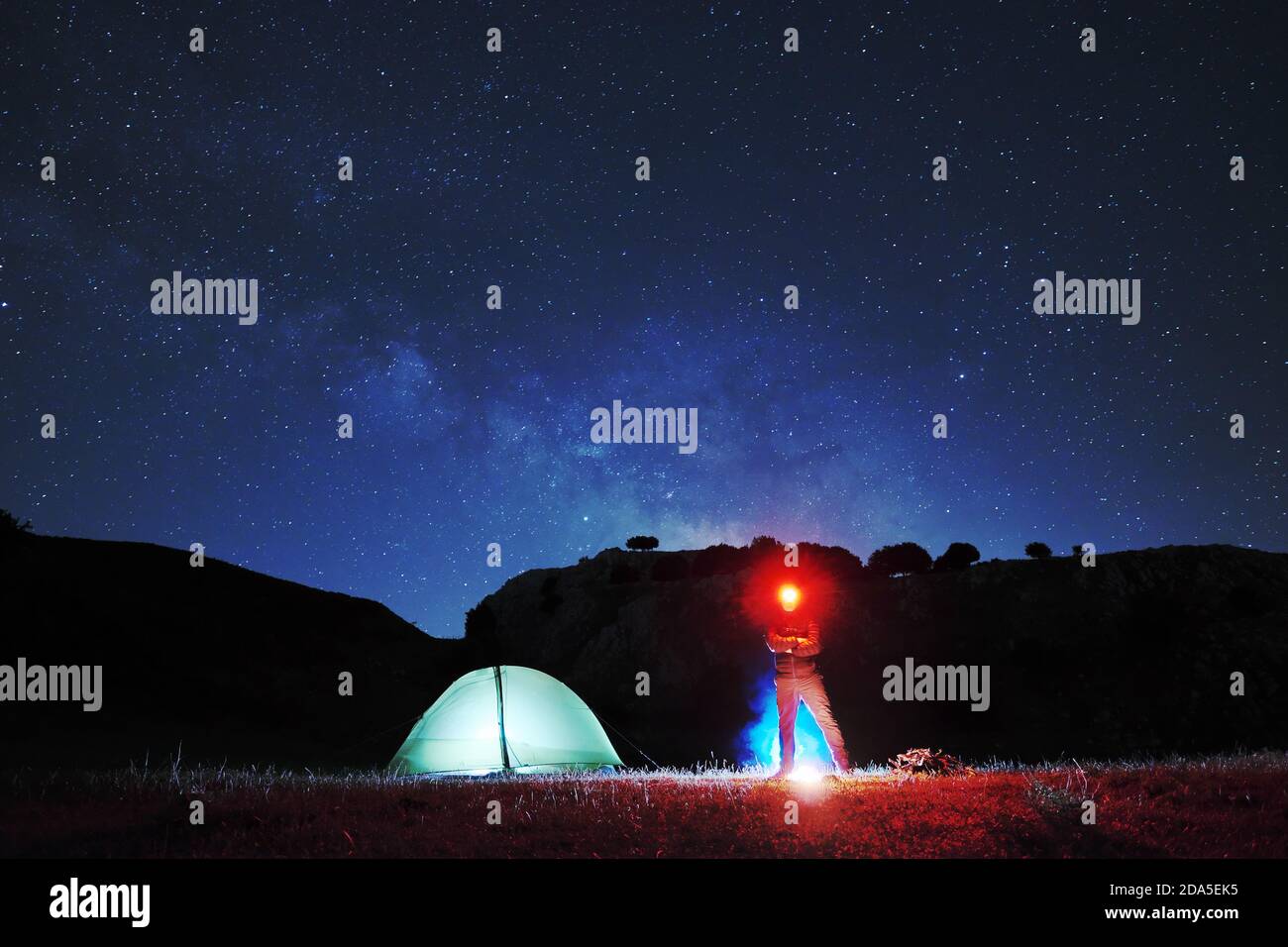 Uomo con faro rosso che guarda in telecamera vicino alla tenda di illuminazione, sullo sfondo cielo stellato sopra la montagna del Parco Nebrodi Foto Stock
