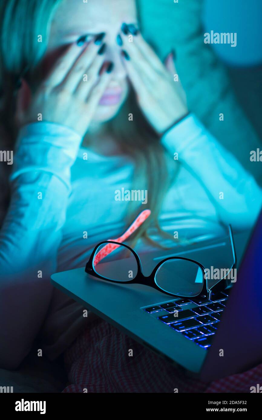 Giovane donna con gli occhiali che le sfregano gli occhi, si sente stanca dopo aver lavorato su un computer portatile, sdraiato in letto sul cuscino. Lavoro eccessivo, esausto, affaticamento. Foto Stock