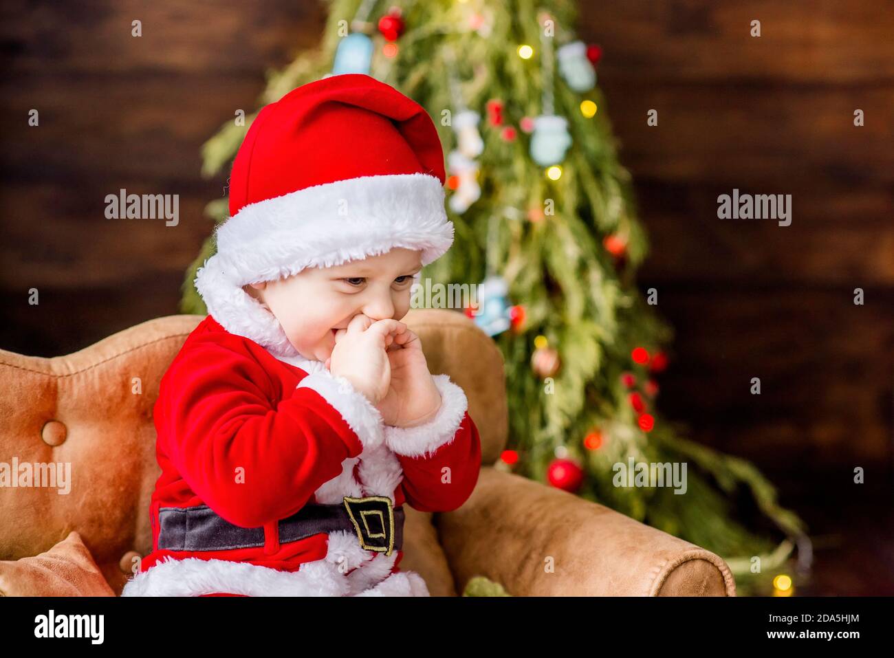 Un bambino piccolo in costume rosso di Babbo Natale, gioca nella sua residenza, lecca una palla di Capodanno come caramella. Cartolina festiva. Il ragazzo biondo ride. Natale Foto Stock
