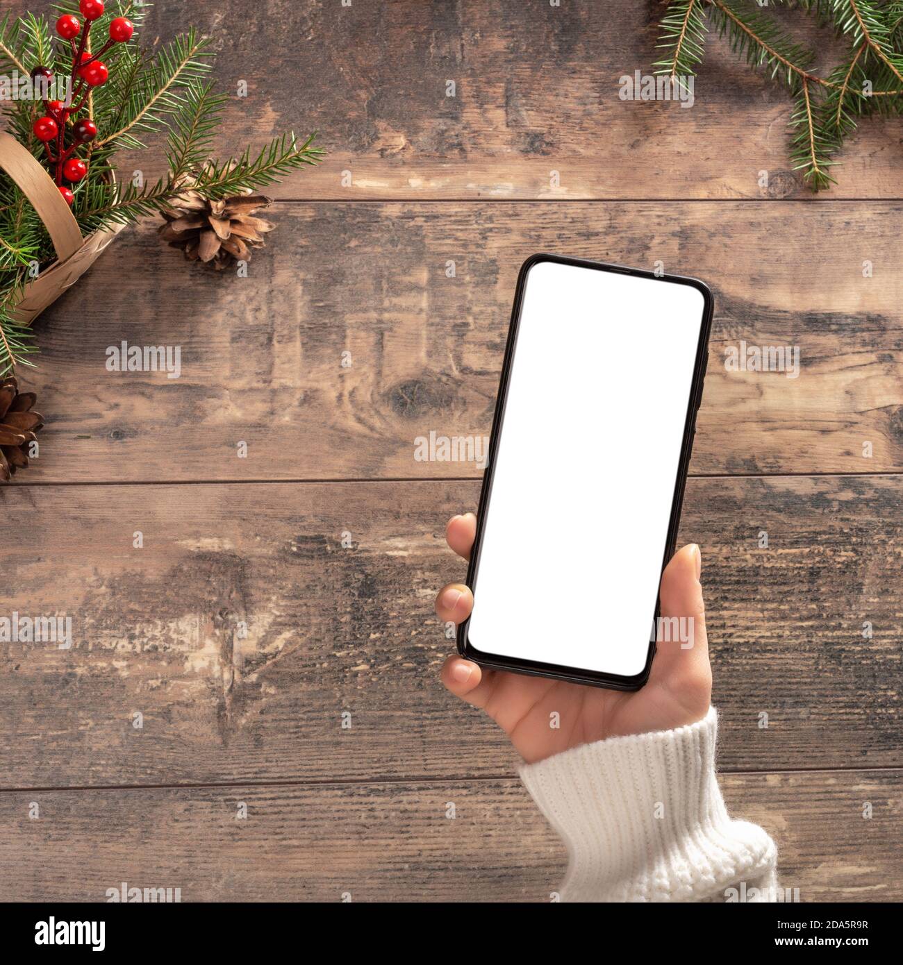 Mockup cellulare su sfondo tavolo di legno durante le vacanze di Natale. Donna tiene un telefono cellulare con uno schermo bianco vuoto Foto Stock