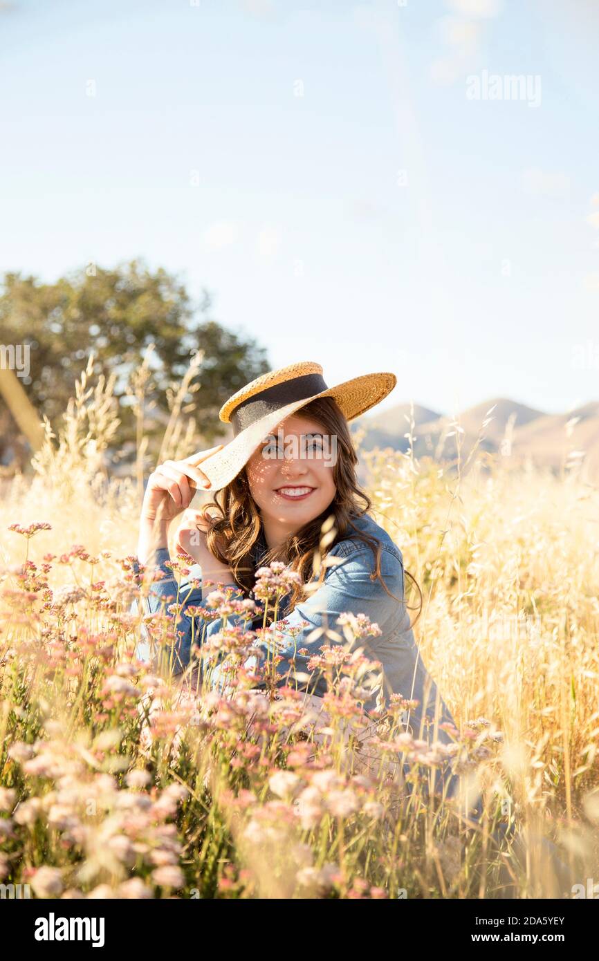 Bella giovane donna sorridente in jeans vestito e cappello di paglia seduto in un prato di fiori rosa e di erbe dorate Foto Stock