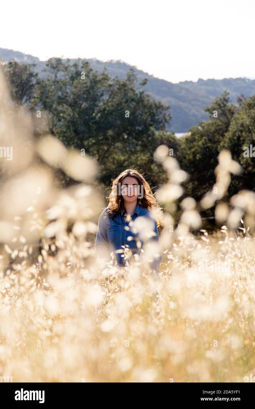 Bella giovane donna con capelli marroni e pelle chiara vista a distanza, in piedi in un prato di bosco di quercia attraverso un bokeh di erbe dorate Foto Stock