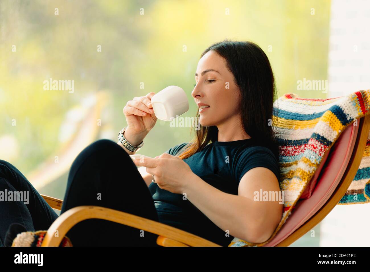 Una giovane bruna sorridente che si gode di una splendida giornata seduta su una sedia a dondolo con una tazza del suo caffè preferito. Foto su sfondo sfocato. Foto Stock