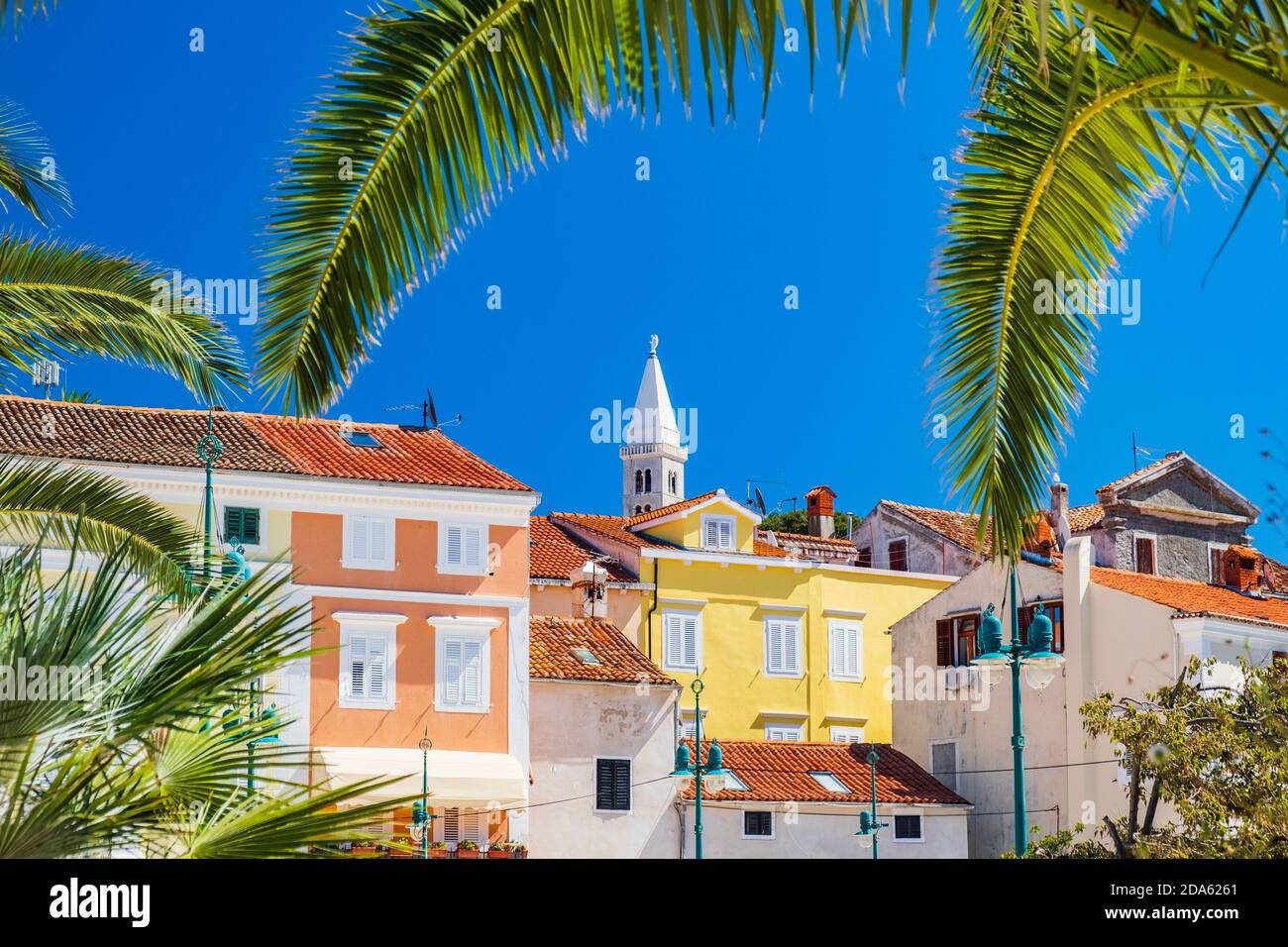 Bellissima città di Lussino sull'isola di Lussino, la costa adriatica in Croazia, la torre della cattedrale e il centro della città, vista attraverso le foglie di palma Foto Stock