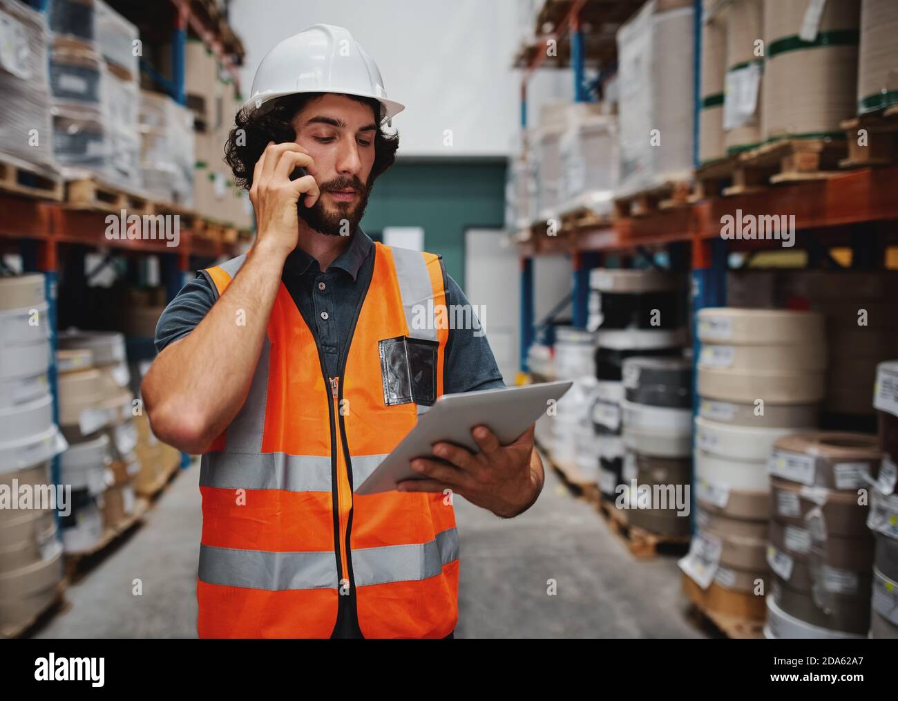 Il responsabile del magazzino tiene traccia dei dettagli degli ordini di fornitura utilizzando un tablet digitale mentre è in conversazione sul telefono cellulare in magazzino Foto Stock