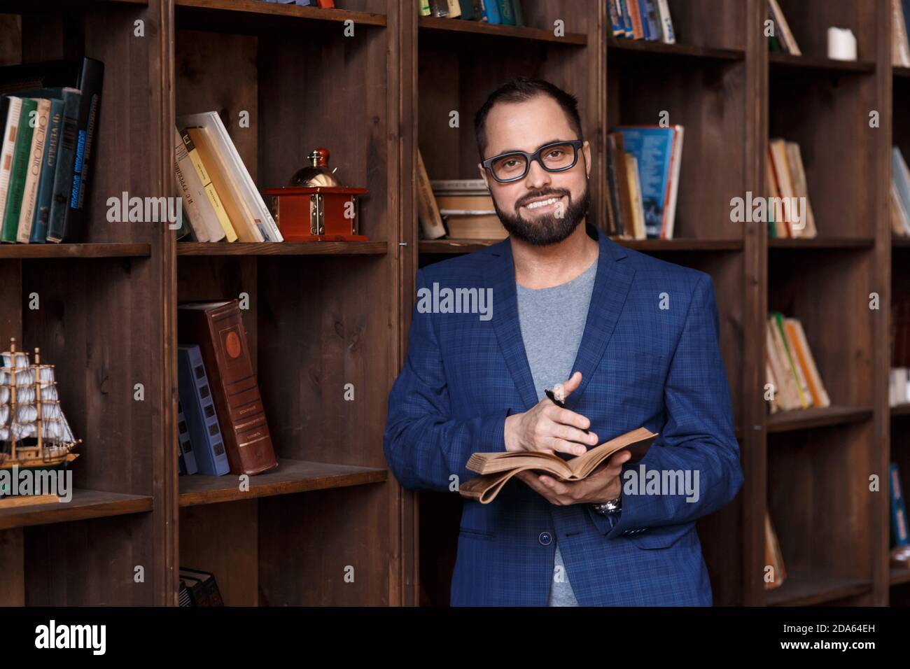 uomo d'affari bearded con gli occhiali in biblioteca con un taccuino in mano sorride. Giovane scrittore Foto Stock