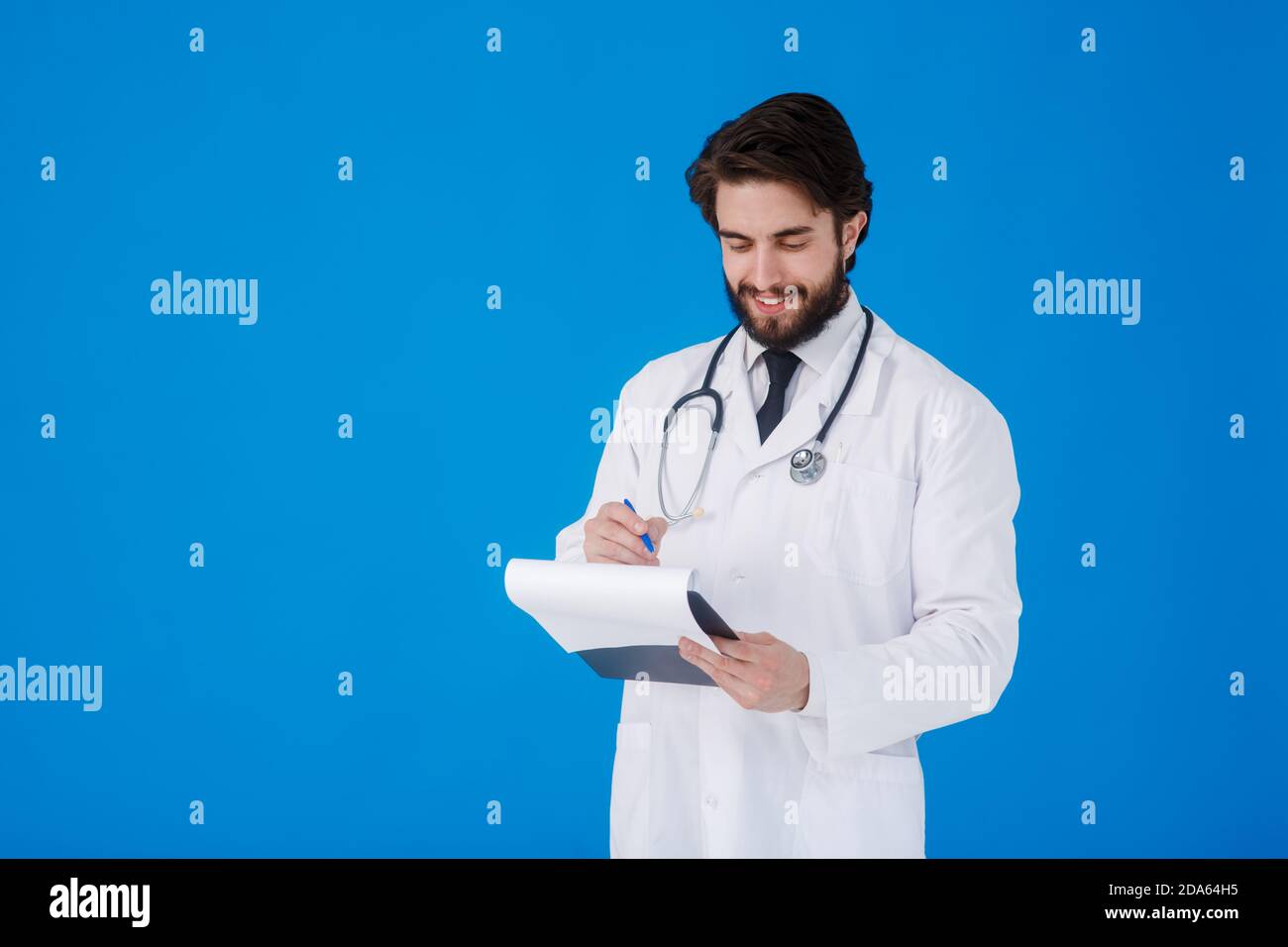 un giovane medico con la bearded in un cappotto medico bianco sopra uno sfondo blu scrive i reclami del paziente su un tablet con documenti e medici Foto Stock