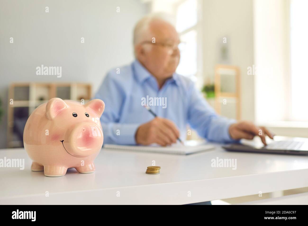 Piggy banca in piedi sulla scrivania con l'uomo in pensione sfocato utilizzando laptop e fare contabilità in background Foto Stock