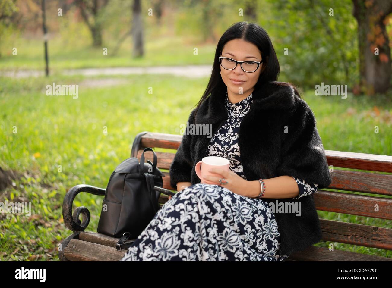 La donna sorridente tiene la tazza di caffè che si rilassa all'aperto nelle giornate di sole. Bella donna asiatica che indossa un cappotto di pelliccia con una tazza di bevanda calda riposante sulla panchina in città Foto Stock