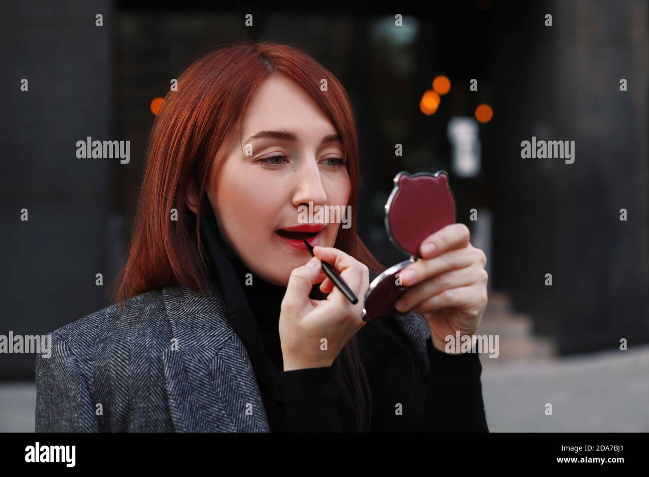 Donna caucasica d'affari rossa guardando piccolo specchio tasca e. applicare il rossetto rosso dopo il lavoro Foto Stock