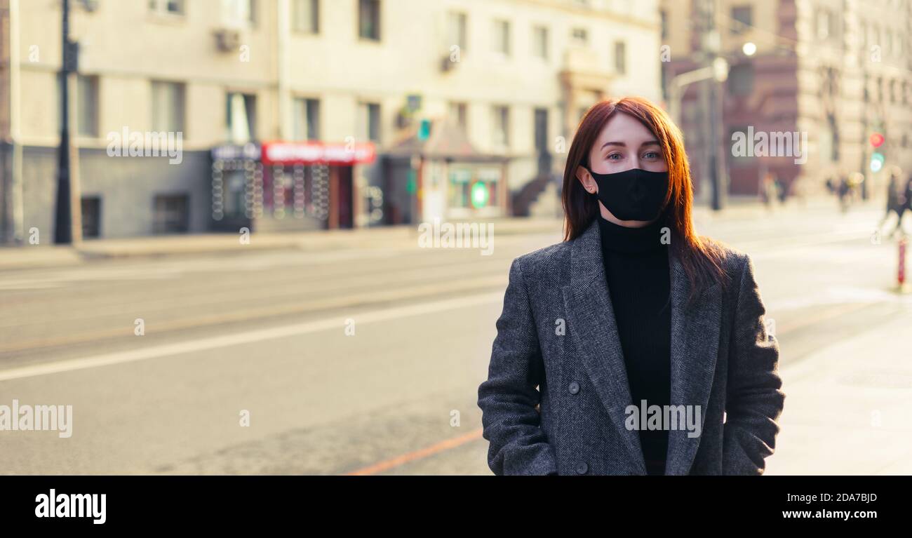 Donna rossa in camice grigio e maschera protettiva nera in piedi su una strada vuota e soleggiata. Foto Stock