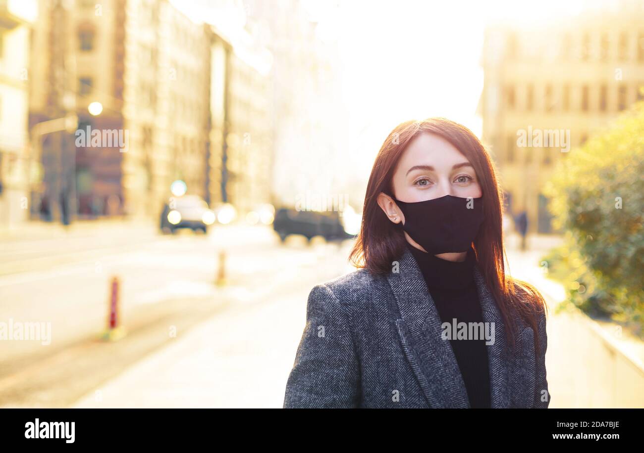 Donna caucasica rossa su una strada vuota luminosa e soleggiata in maschera protettiva nera. Rinascita dopo il coronavirus. Foto Stock