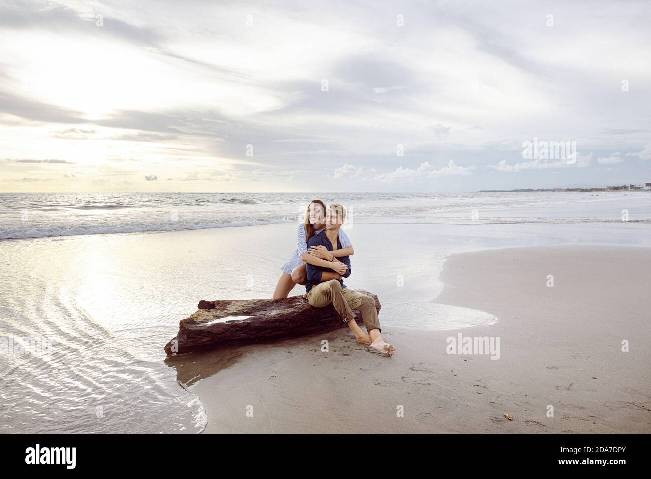 Coppia seduta su un tronco di albero morto su una spiaggia a Bali al tramonto, guardando lontano Foto Stock