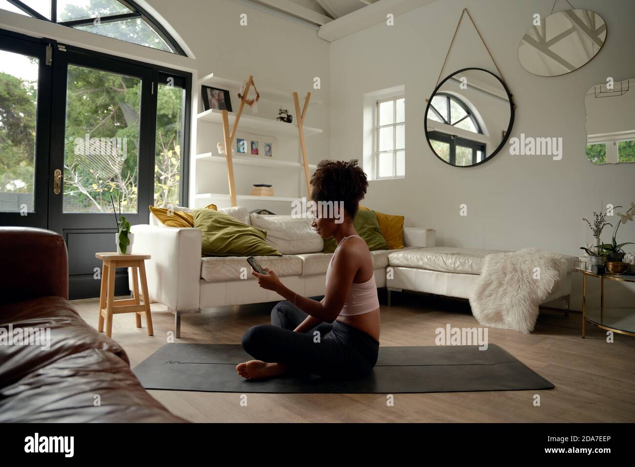 Giovane donna africana che usa il cellulare mentre si siede sul tappeto di yoga a casa Foto Stock