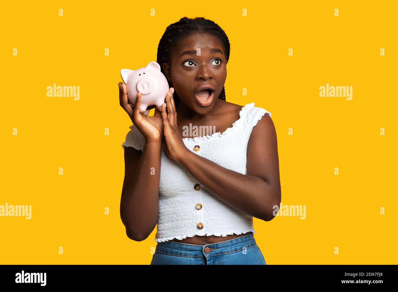 Scioccata donna afroamericana che ascolta il suono delle monete in piggy banca Foto Stock
