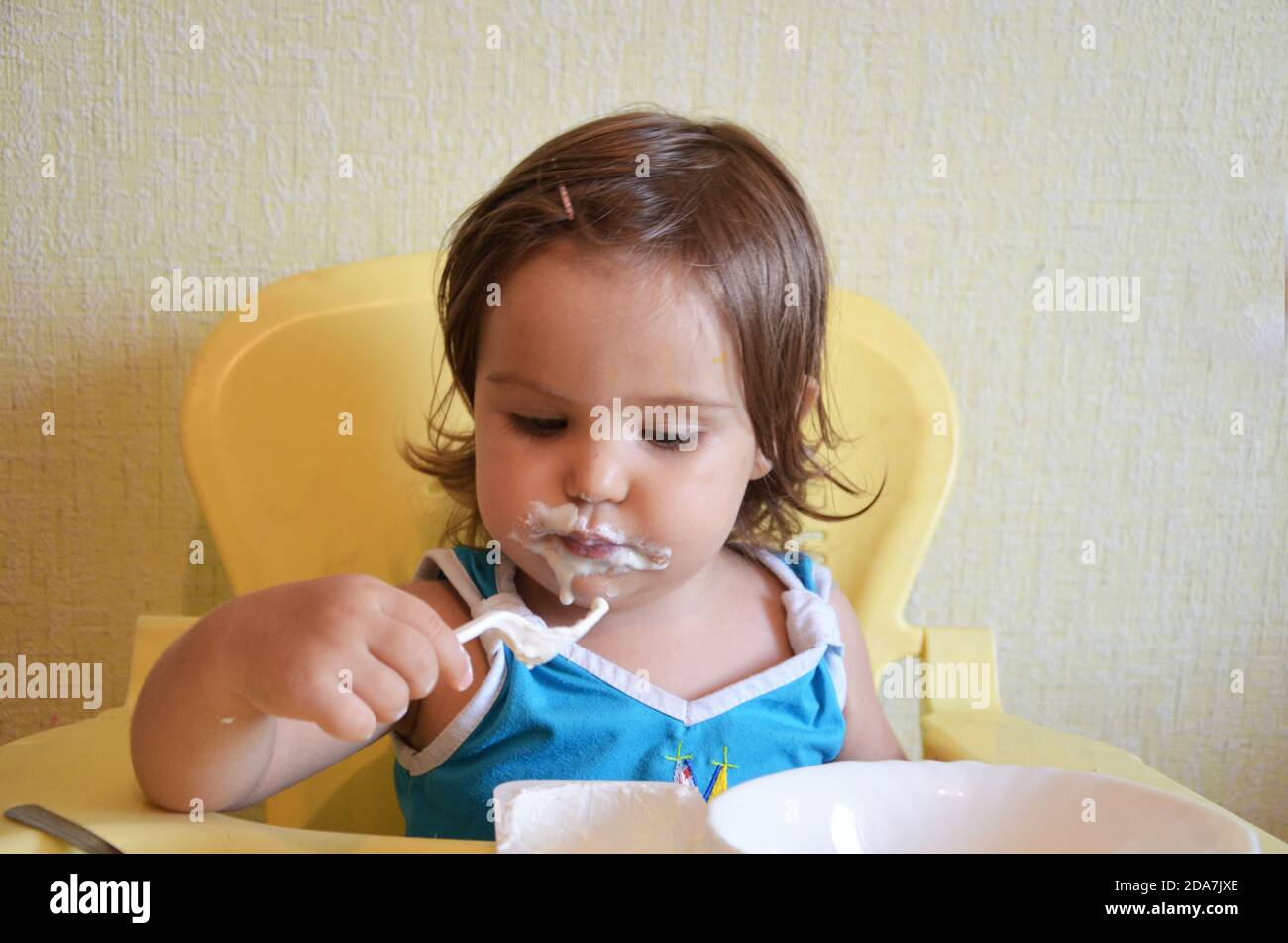 Un bambino piccolo da solo mangia l'alimento del bambino con un cucchiaio da un piattino che si spillano in porridge. Una ragazza o due, mangia con un cucchiaio di plastica o forchetta formaggio cottage Foto Stock