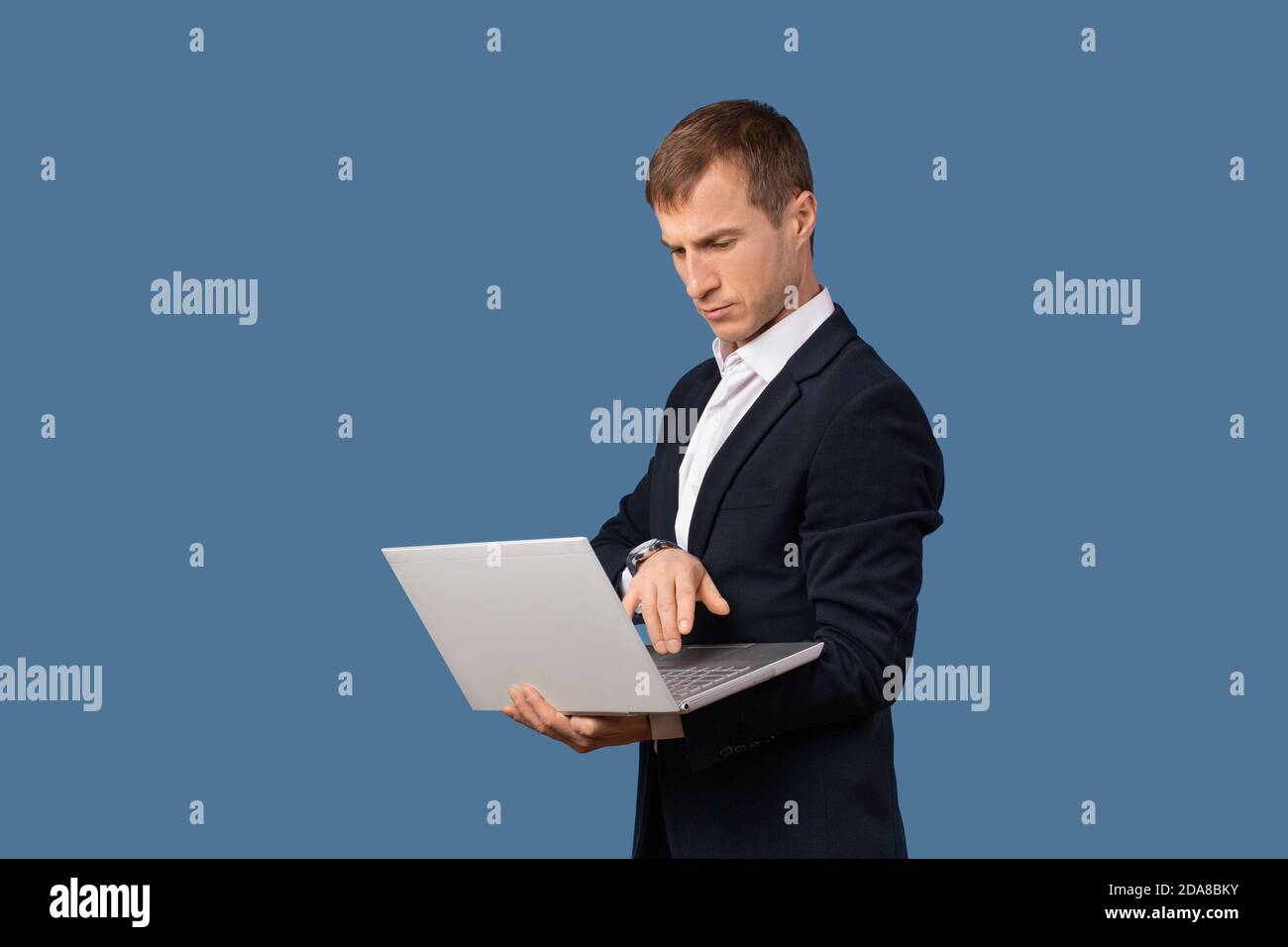Un uomo d'affari in un vestito da lavoro guarda un laptop sul peso nelle sue mani. Studio shot concetto di lavoro remoto senza ufficio su sfondo blu Foto Stock