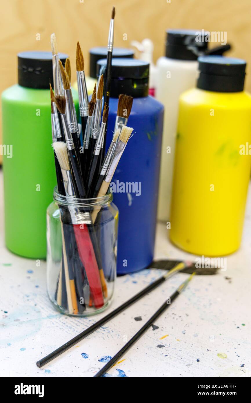 Primo piano di uno studio d'arte o di uno spazio di artisti, l'immagine mostra pennelli di vernice in una tazza di vetro con bottiglie di colori acrilici Foto Stock