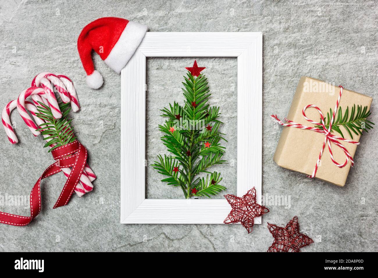 Albero di Natale minimalista fatto di sempreverde pianta in bianco cornice foto, dono e canne caramelle avvolto in nastro rosso. Composizione creativa piatta. Vinci Foto Stock